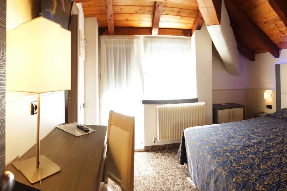 Een hotelkamer met een houten plafond, een bed met een bedsprei met dessin, een bureau met een lamp en een radiator.