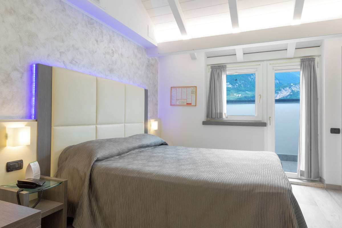 Een hotelkamer met een bed, nachtkastje, lamp en uitzicht op een waterpartij door een raam.