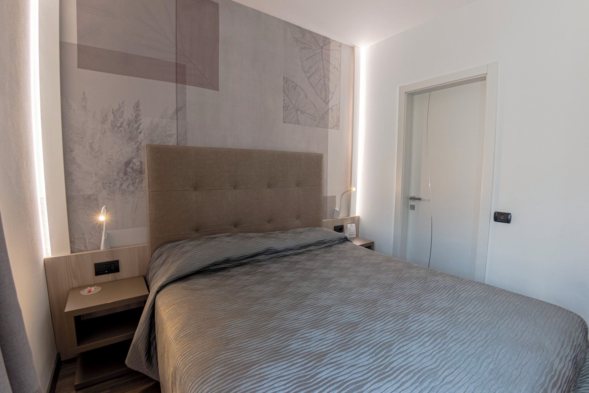 A bedroom with a bed, bedside tables, a nightstand, a wall-mounted lamp, and a door.