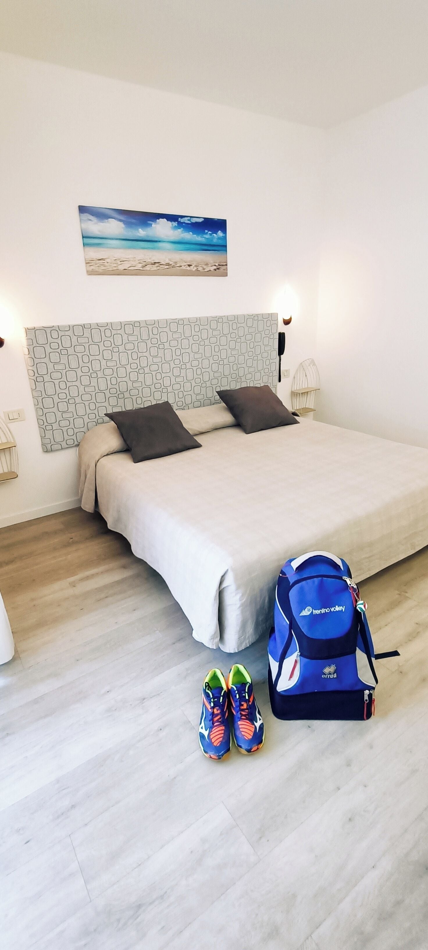 Un dormitorio con una cama, cabecera con estampado geométrico, mesita de noche, lámpara, teléfono instalado en la pared, cuadro de una playa, una mochila azul y un par de zapatillas de colores.