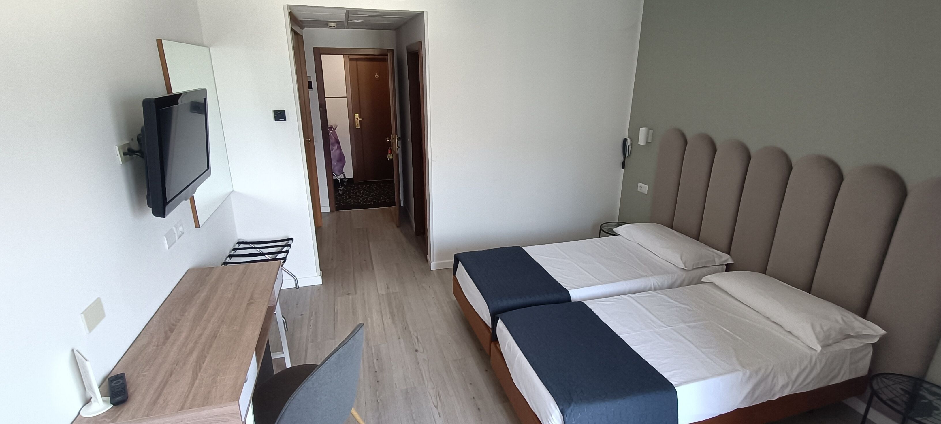 A hotel room with a bed, a desk, a chair, a TV, and a door leading to a hallway.