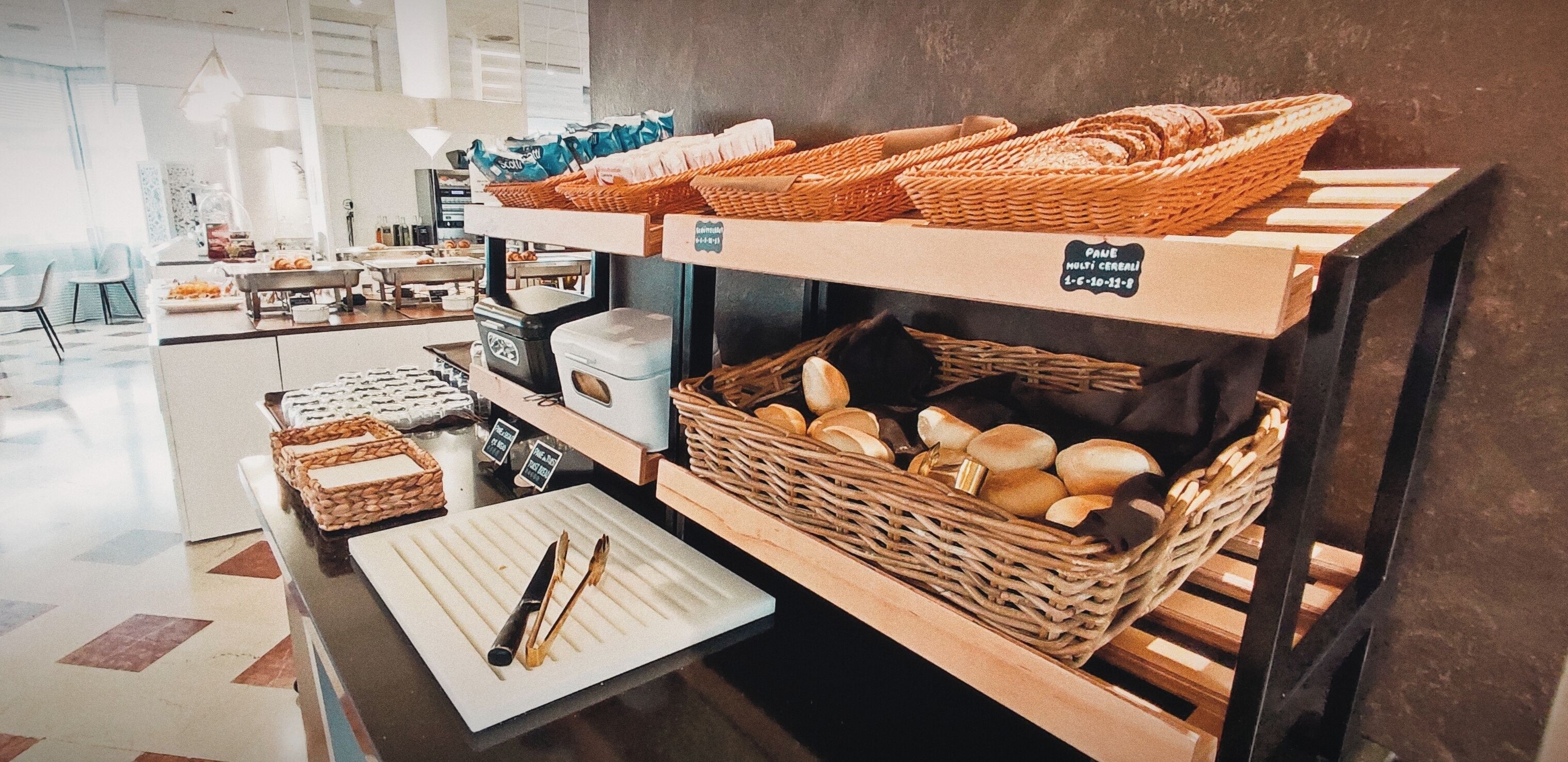 A bakery display with bread and pastries in wicker baskets, a sign indicating 'PAJE HOLSTI CEREALI', and a clean, modern interior with a tiled floor.