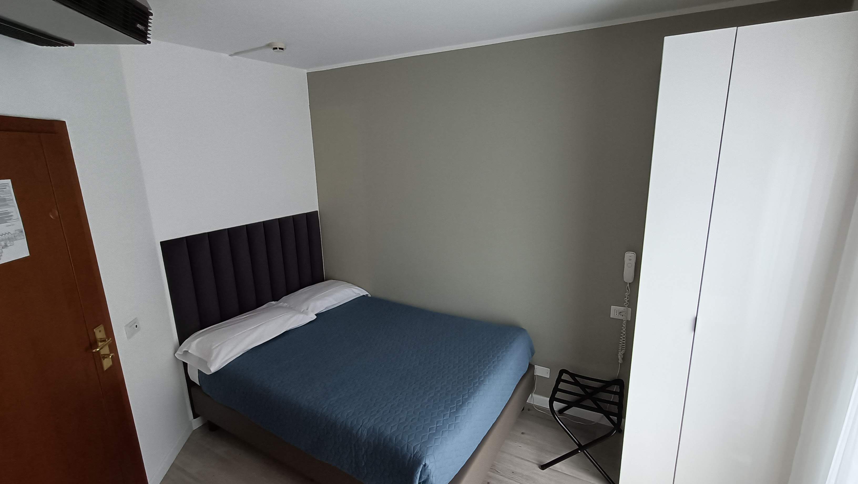 Une petite chambre avec un lit simple, dotée d’une tête de lit sombre, d’une couvre-lit bleue, d’une armoire et d’une petite table avec une chaise.