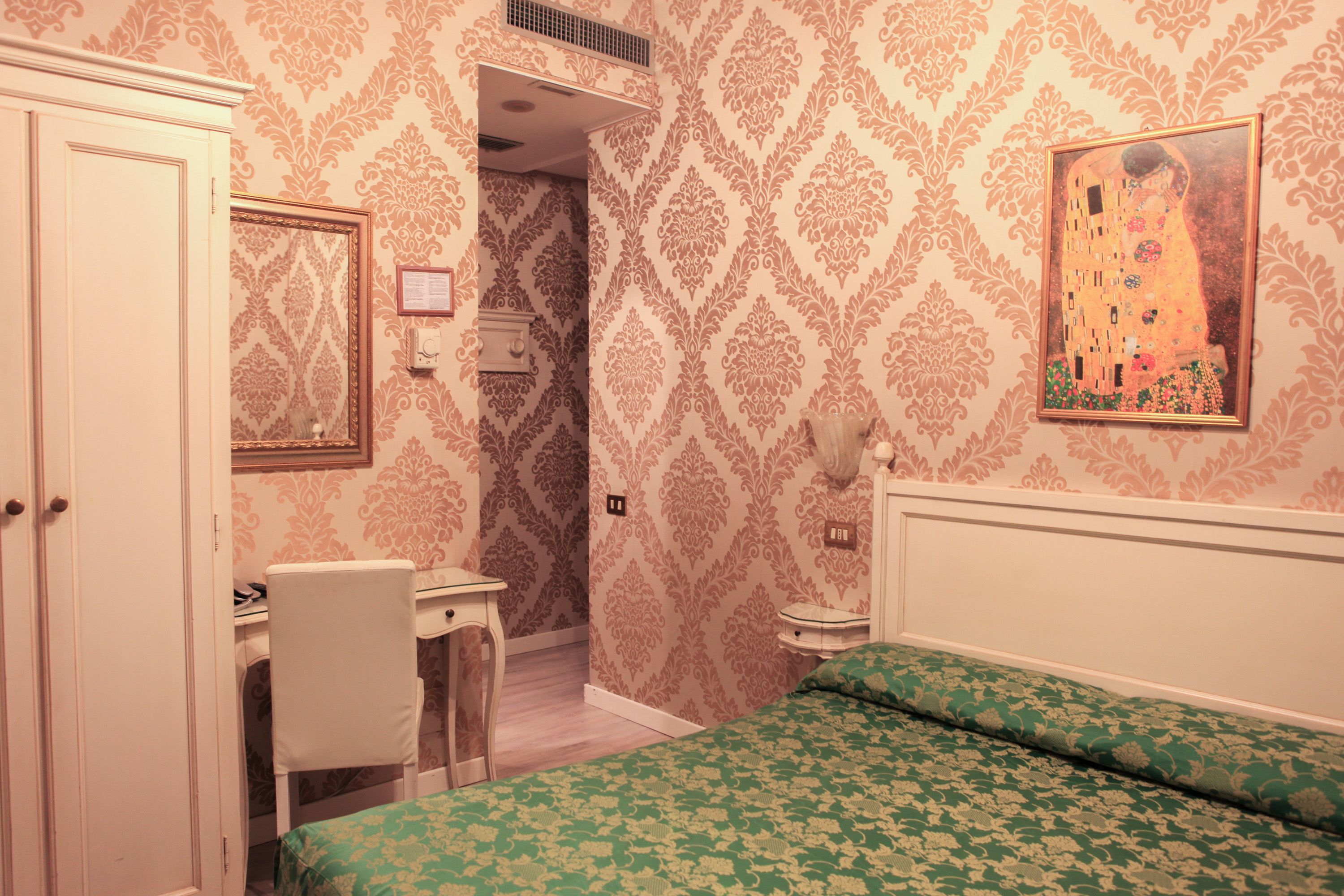 A bedroom with a bed, a desk, and a chair. There is a painting on the wall and a door.