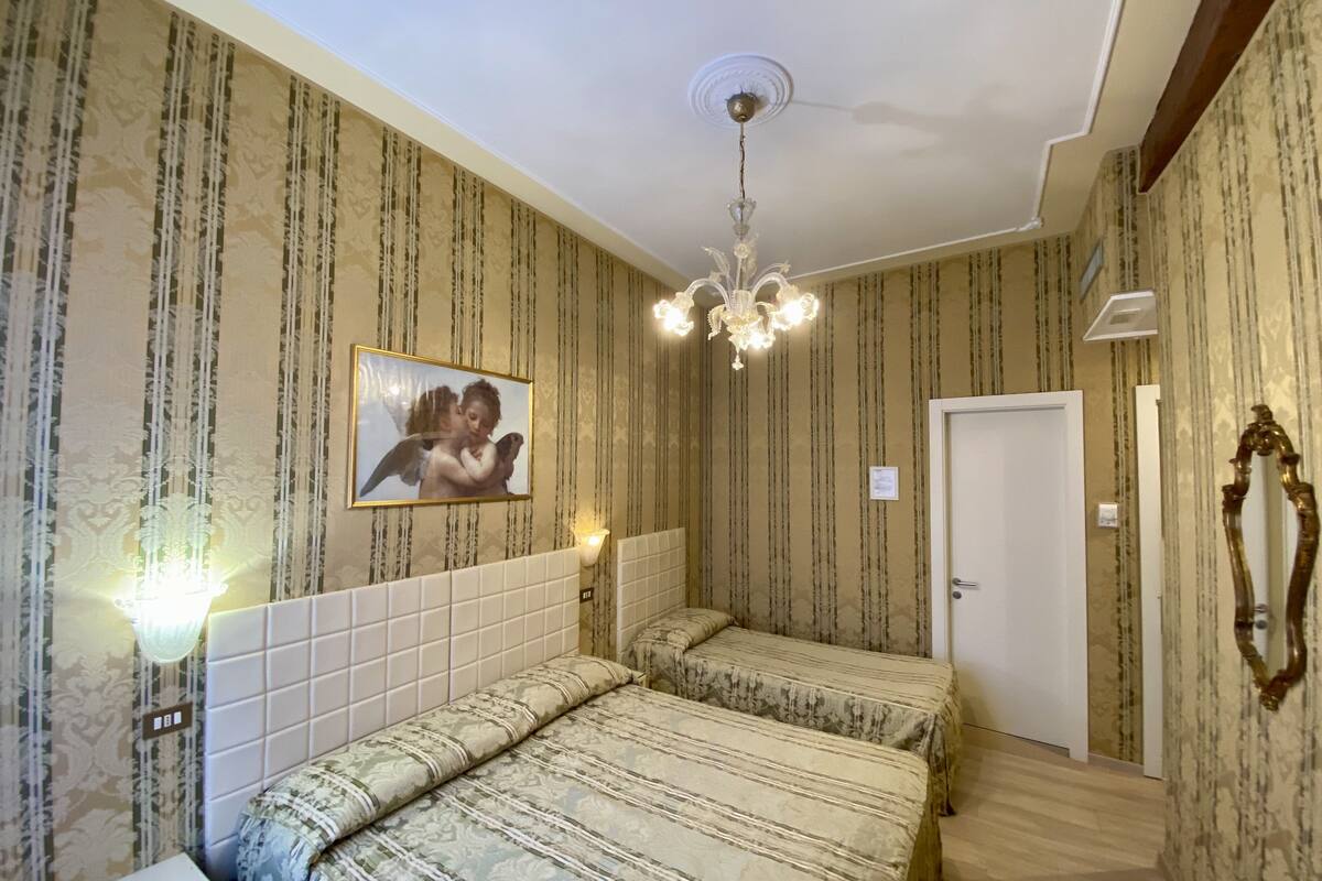 Hotellihuone, jossa on kaksi sänkyä, kattokruunu, seinällä oleva taulu ja ovi.