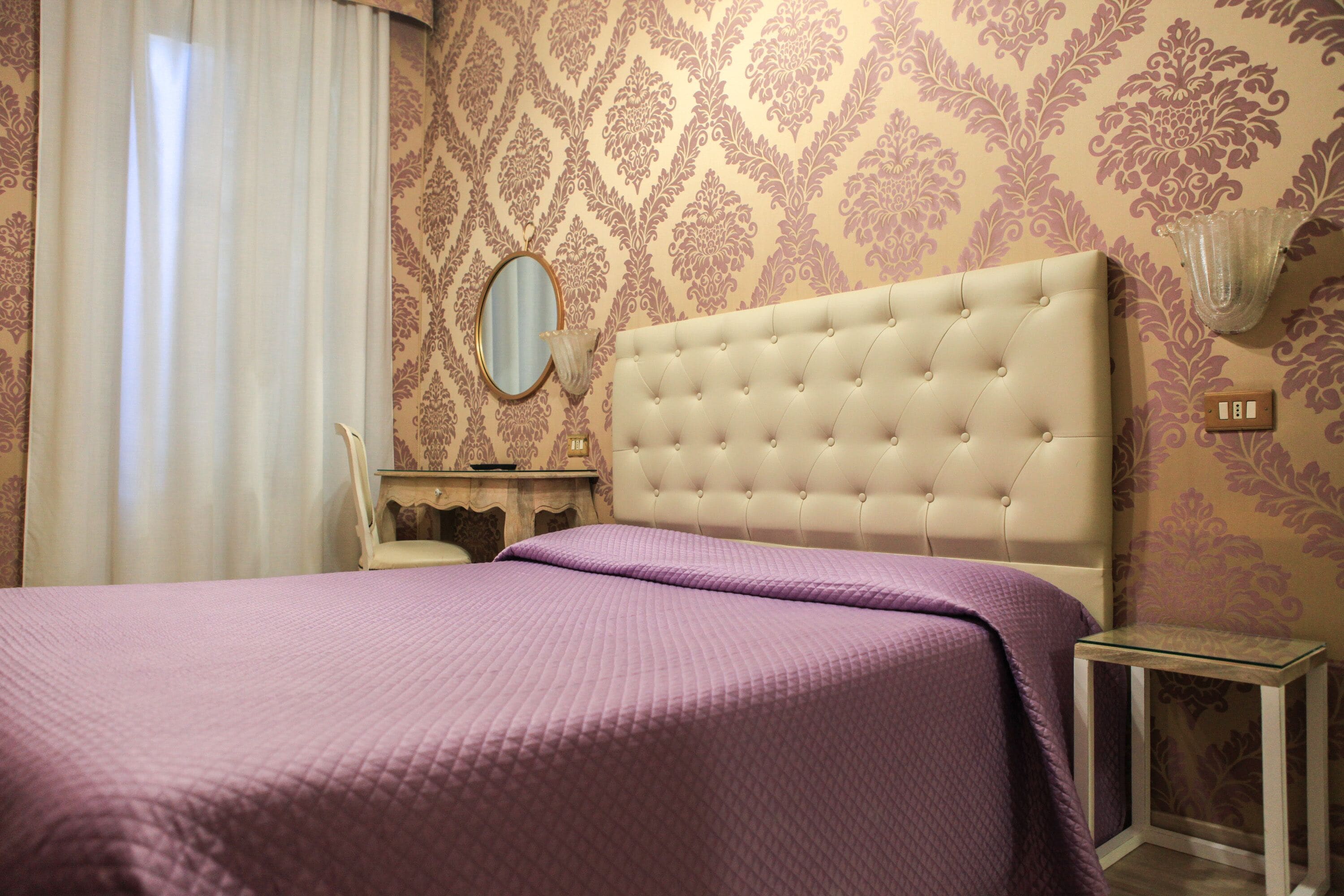 A bedroom with a tufted headboard, a bed with a purple bedspread, a bedside table, a mirror, and patterned wallpaper.