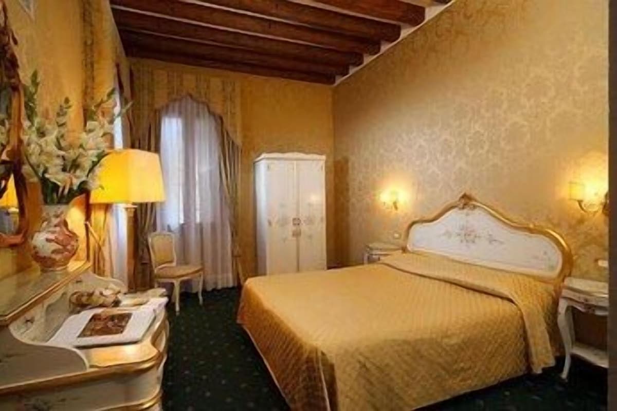 Hotellihuone, jossa on sänky, työpöytä lampun kanssa, tuoli, vaatekaappi ja seinään kiinnitetty valaisin.