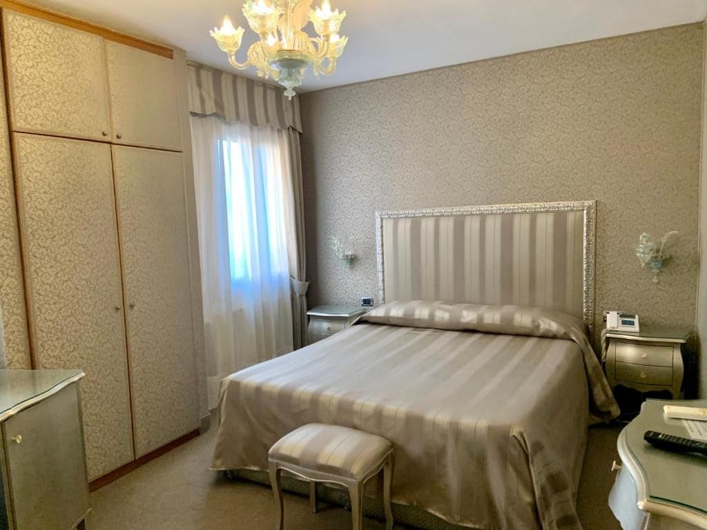 Une chambre d’hôtel avec un grand lit, une table de chevet, une commode, une chaise, une armoire et un lustre.