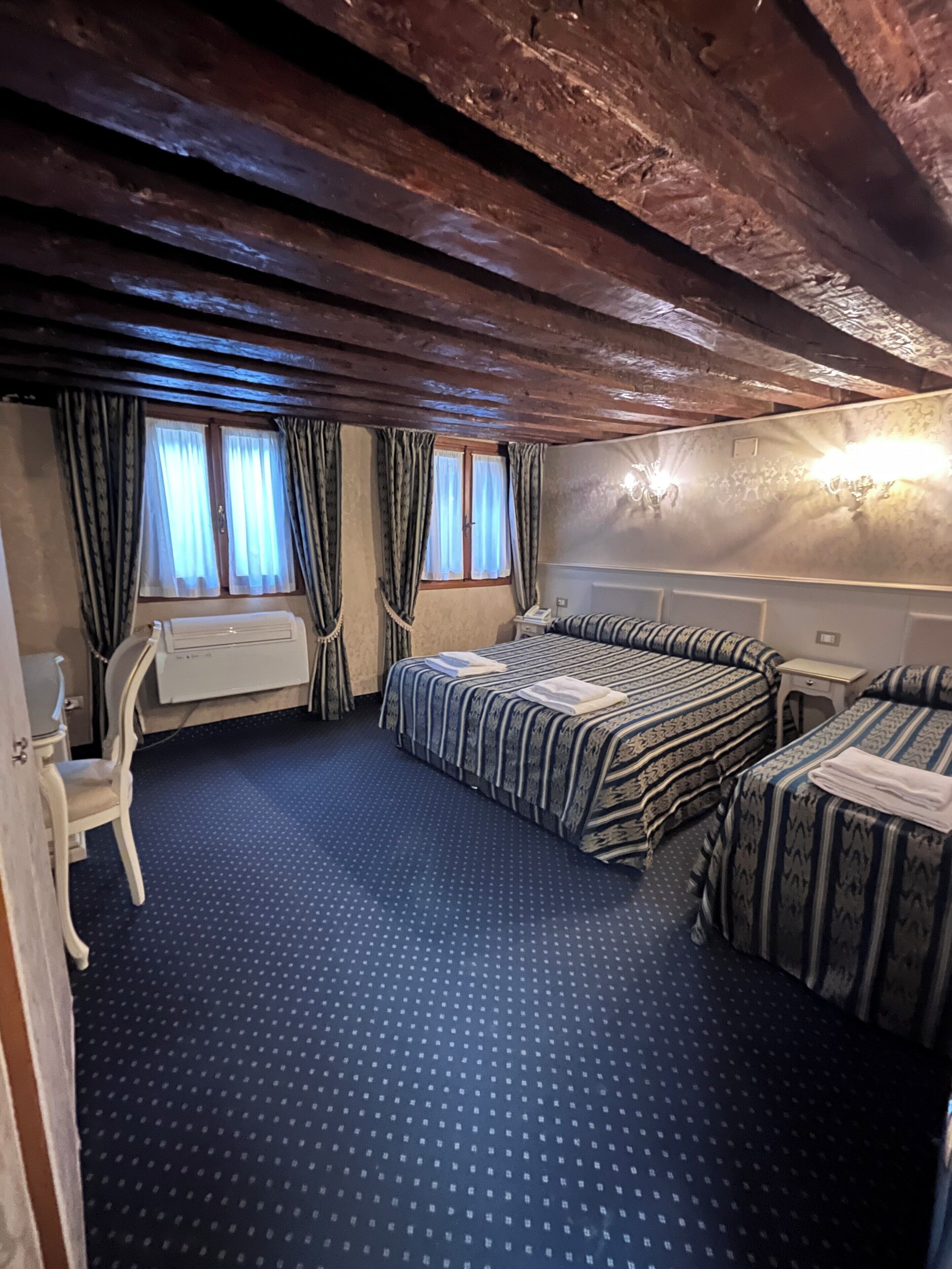 Une chambre avec deux lits, un bureau et une chaise. Les fenêtres sont dotées de rideaux et la chambre a un plafond en bois.