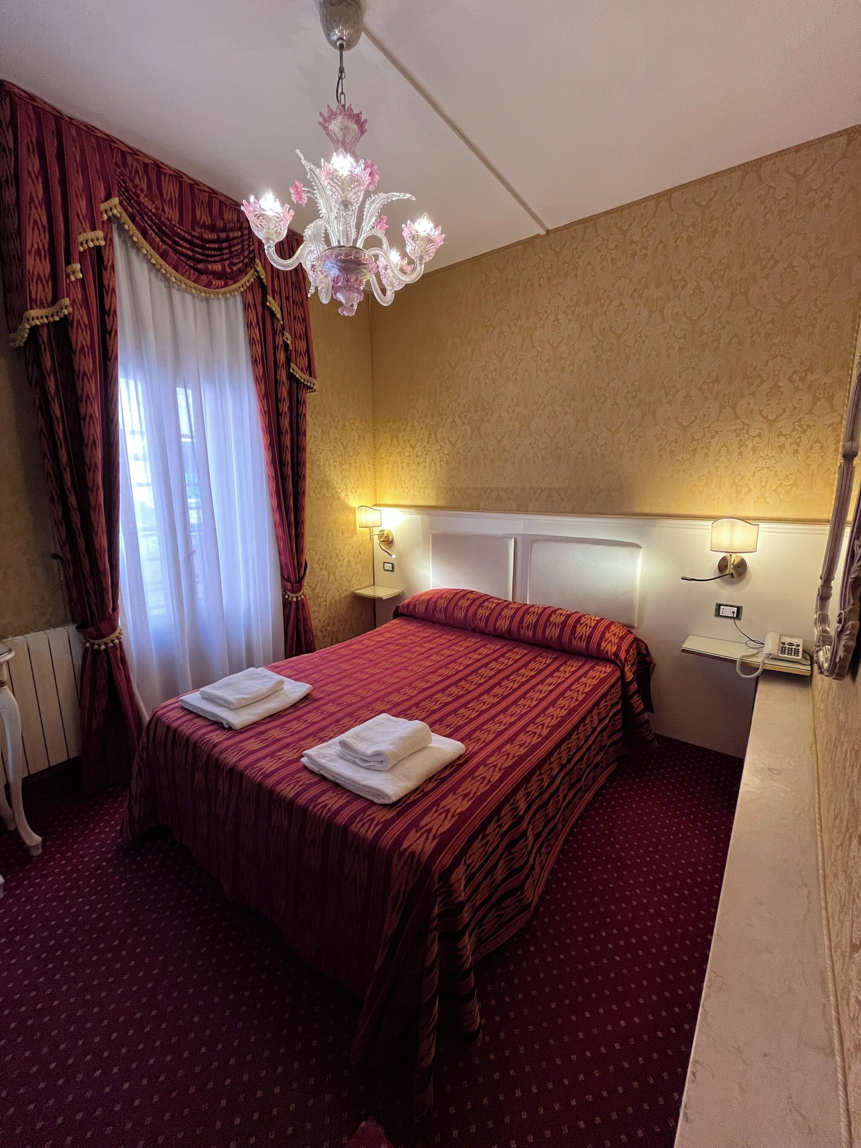 A hotel room with a large bed, two bedside lamps, a chandelier, and a radiator.