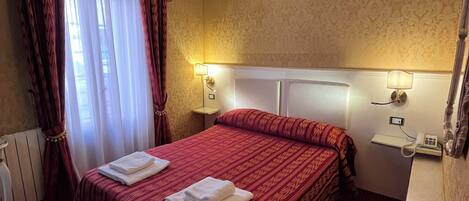 A hotel room with a large bed, two bedside lamps, a chandelier, and a radiator.