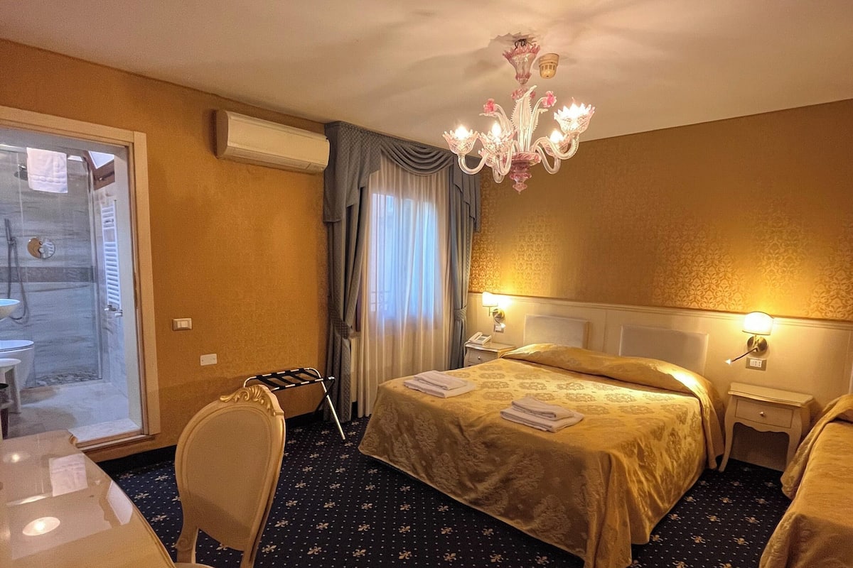 A hotel room with a bed, a desk, a chair, a chandelier, and a window with curtains.