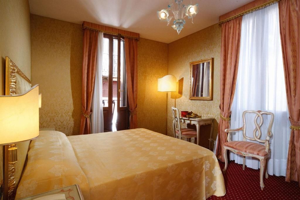 A hotel room with a bed, a desk with a lamp, a chair, a mirror, and curtains.