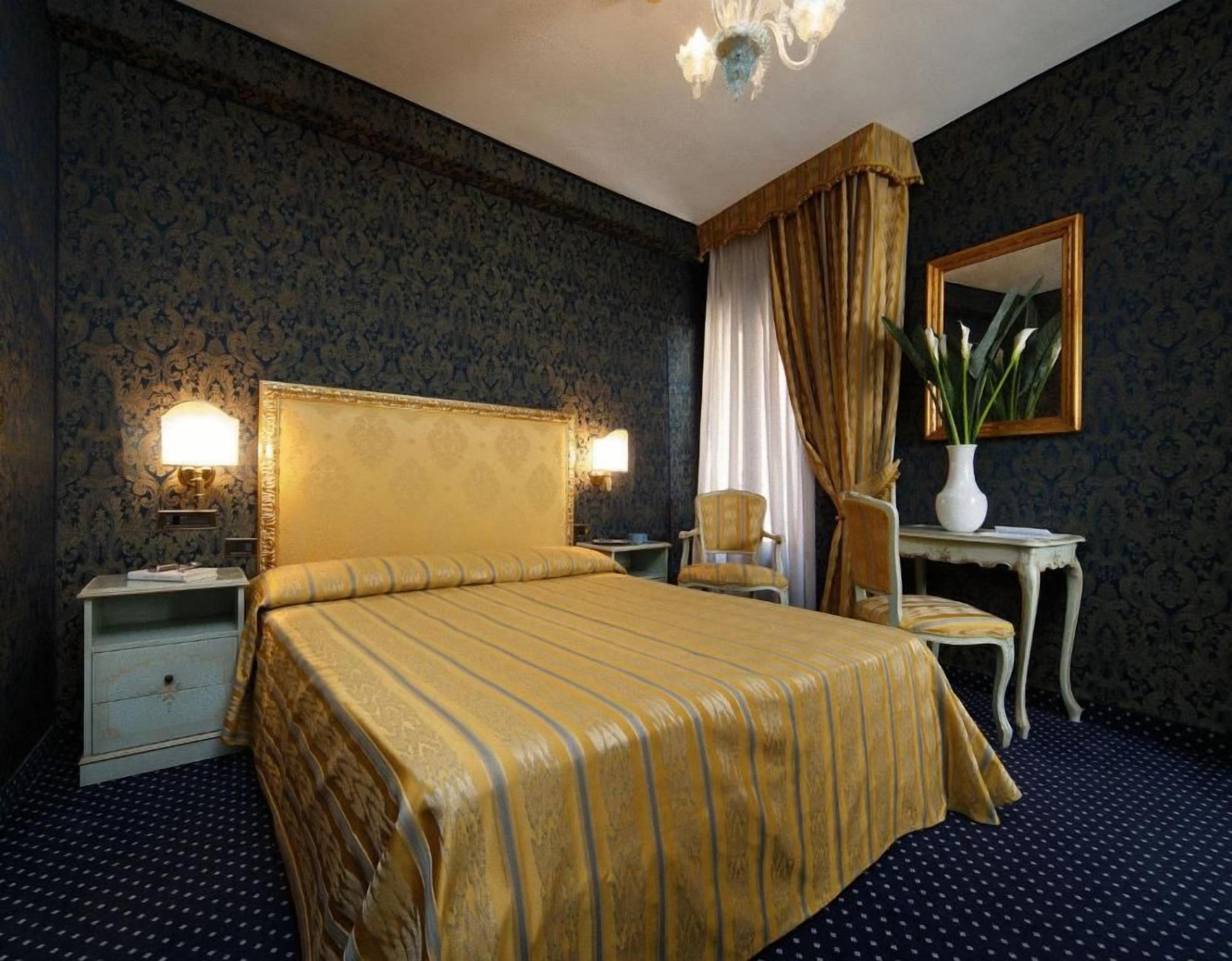 A hotel room with a large bed, a nightstand with a lamp, a chair, a small table with a vase, and a mirror on the wall.