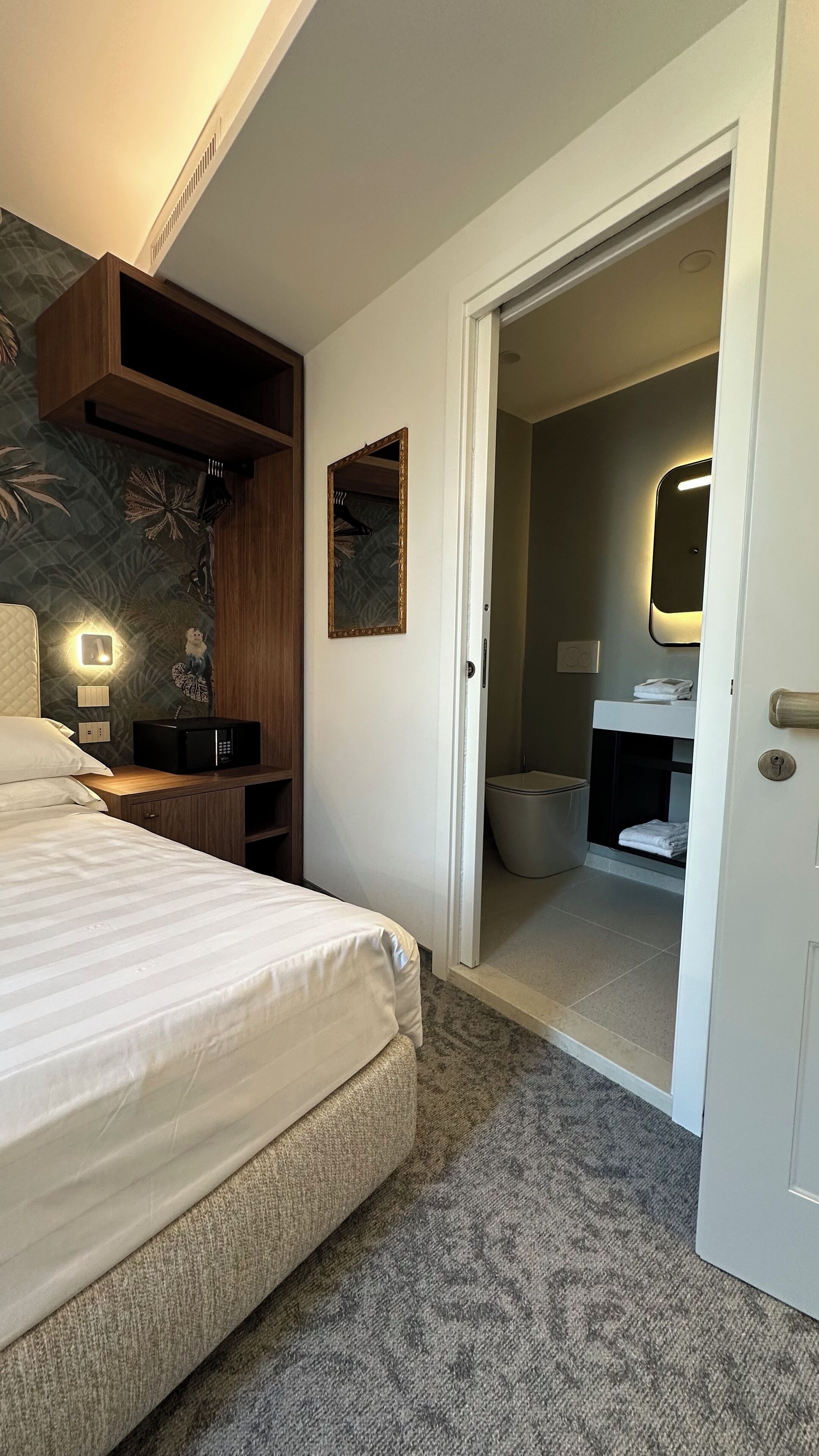 A hotel room with a bed, a wooden shelf, a wall-mounted light, a mirror, and a bathroom visible through an open door.