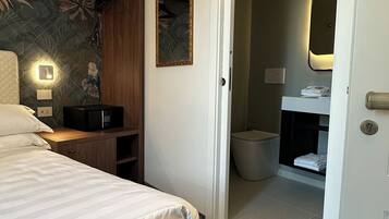 A hotel room with a bed, a wooden shelf, a wall-mounted light, a mirror, and a bathroom visible through an open door.