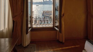 A room with a large window, curtains, a wooden floor, and a view of a river with boats. - Pensione Wildner (Venice)