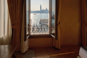 A room with a large window, curtains, a wooden floor, and a view of a river with boats.