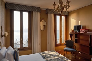 A hotel room with a bed, a chair, a TV, a bookshelf, and large windows with a view of the sea.