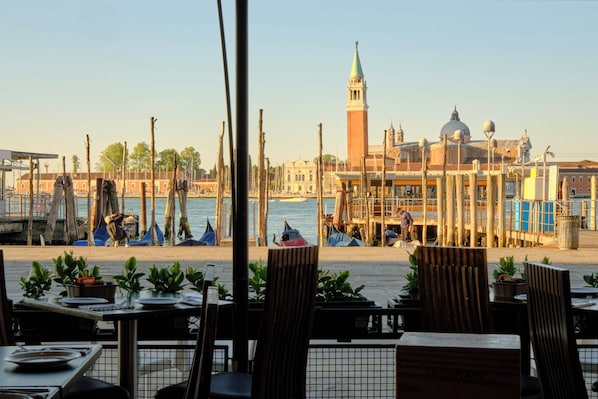 A waterfront restaurant with tables and chairs, a view of the water, and a prominent building with a tower in the background.
