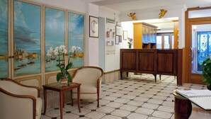 A lobby with a tiled floor, a reception desk, a sofa, a chair, and a painting of a cityscape. - Pensione Wildner (Venice)