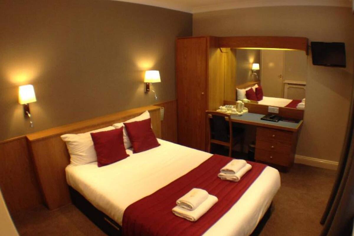 A hotel room with a bed, a desk, a chair, a mirror, and two wall-mounted lamps.