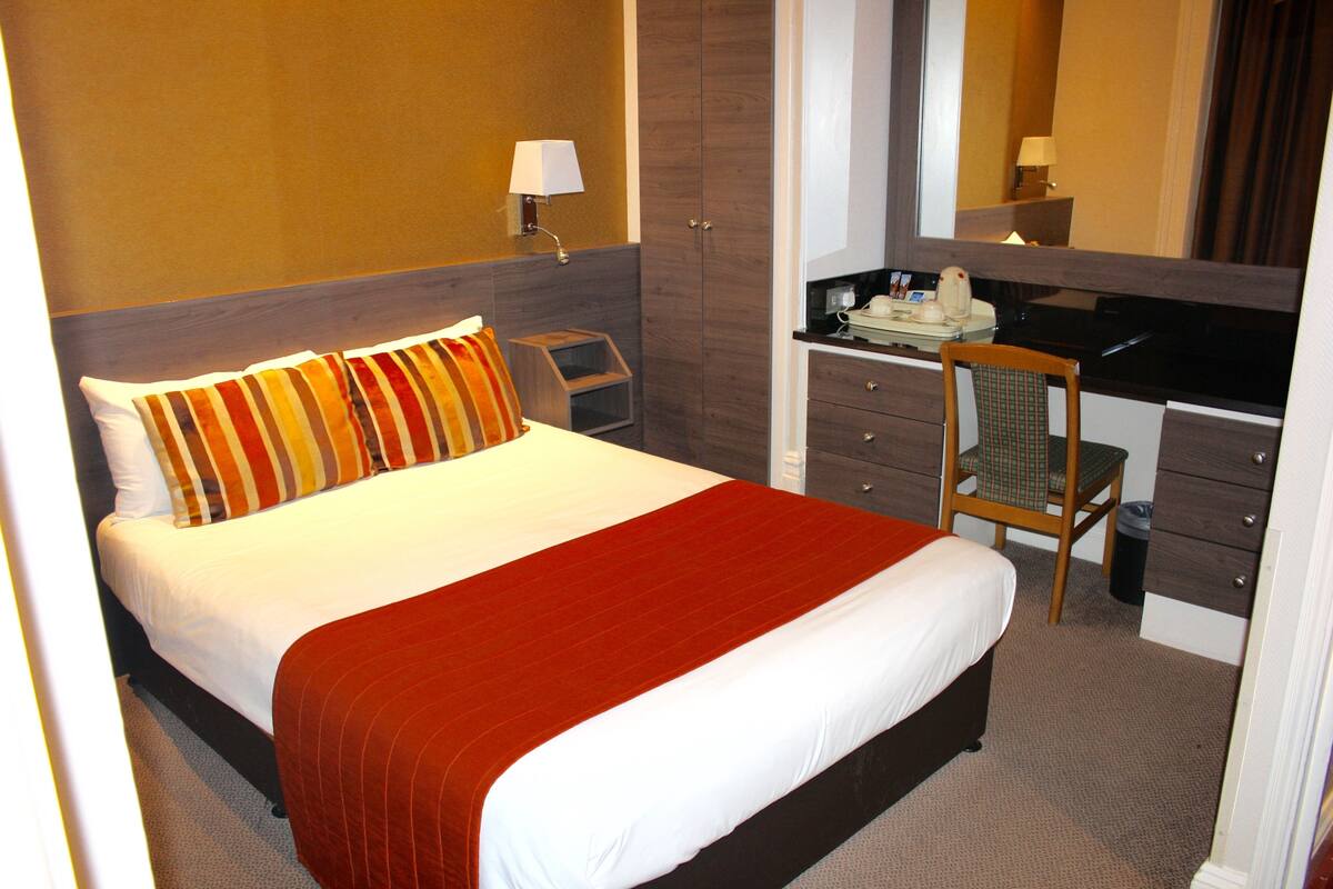 A hotel room with a bed, a desk, a chair, and a trash bin.