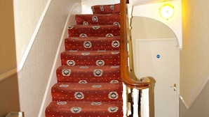 Un escalier recouvert d’une moquette à motifs et doté de rampes en bois, menant à une porte surmontée d’un luminaire.