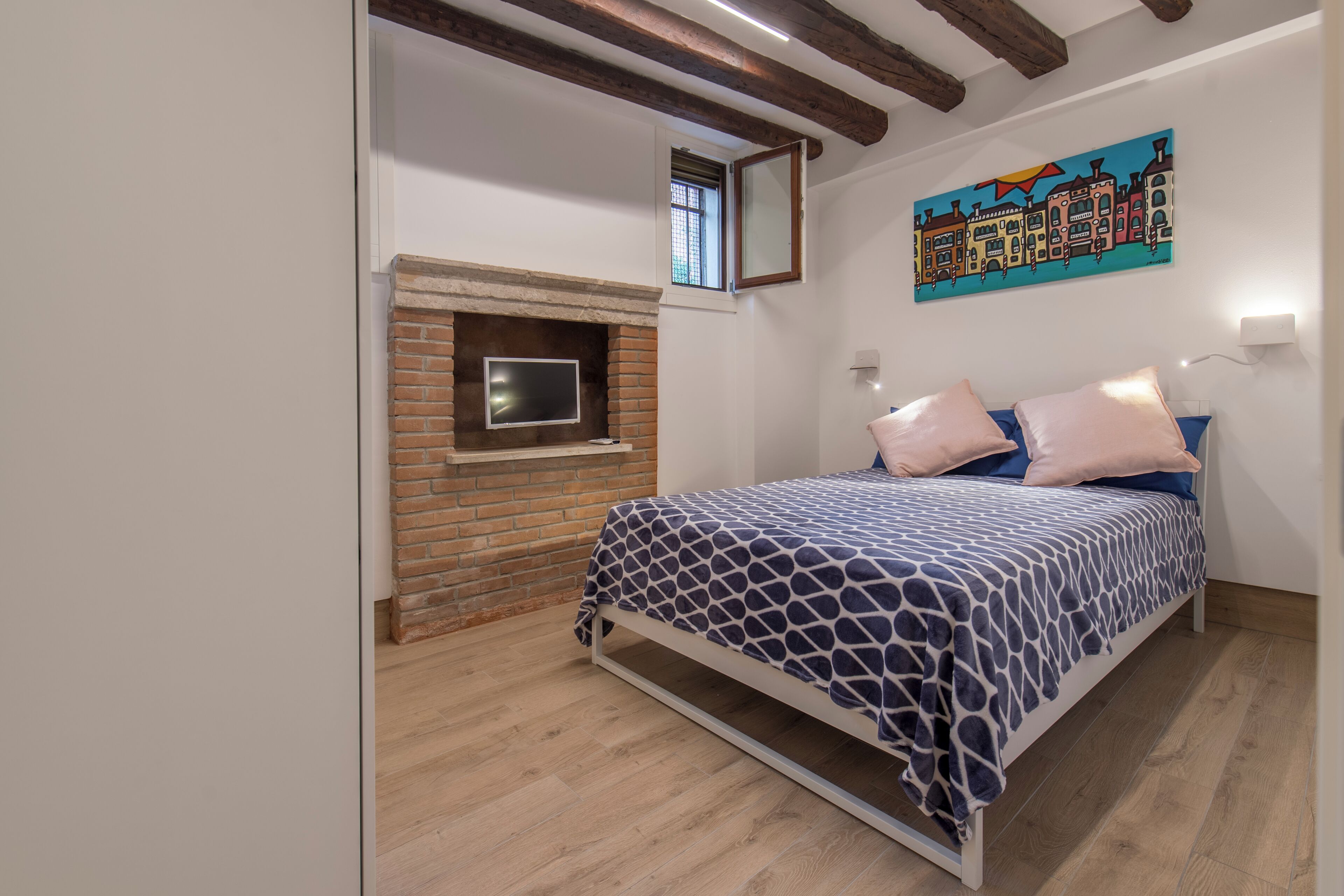 A bedroom with a bed, a brick fireplace, a painting on the wall, and wooden beams on the ceiling.