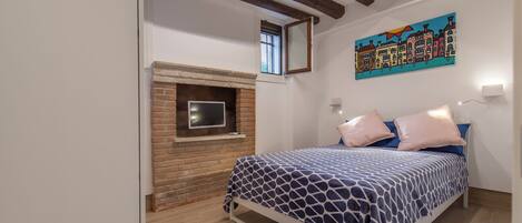 A bedroom with a bed, a brick fireplace, a painting on the wall, and wooden beams on the ceiling.