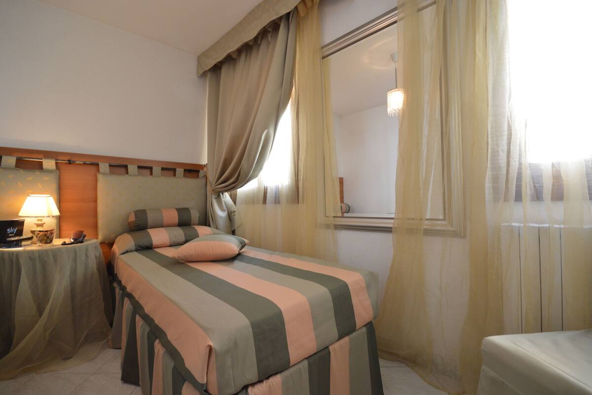 Una habitación de hotel con cama, mesita de noche con lámpara y ventana con cortinas.