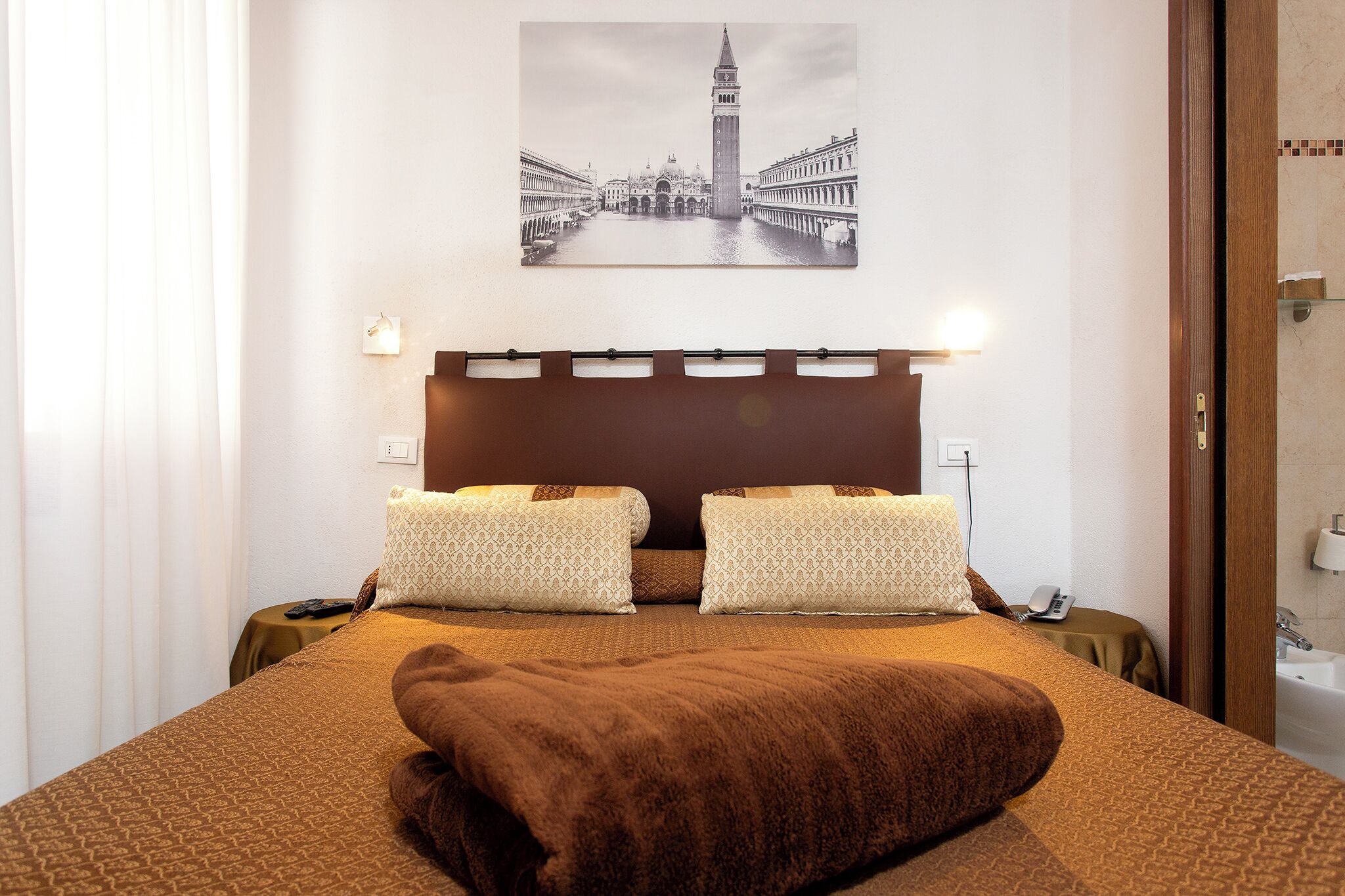 Een hotelkamer met een groot bed, twee kussens, een nachtkastje, een telefoon en een schilderij van een bekende brug en toren aan de muur.