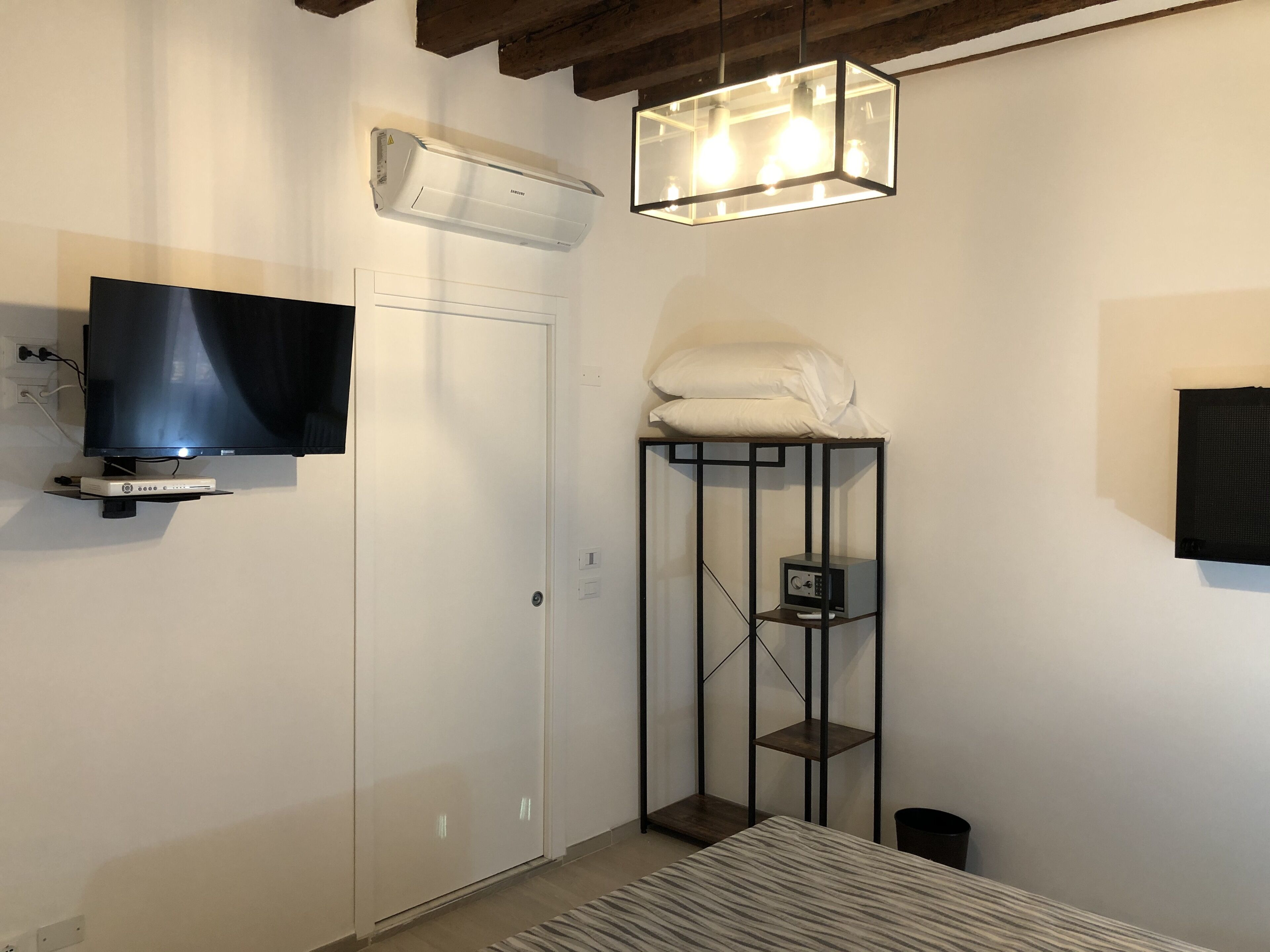 Ett modernt hotellrum med en platt-TV, inbyggd luftkonditionering, en unik lampa, en liten hylla med en radio och en snyggt bäddad säng.