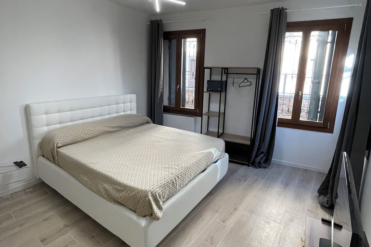 Un dormitorio moderno con una cama blanca, una lámpara de techo, un ventanal con cortinas y un televisor de pantalla plana.