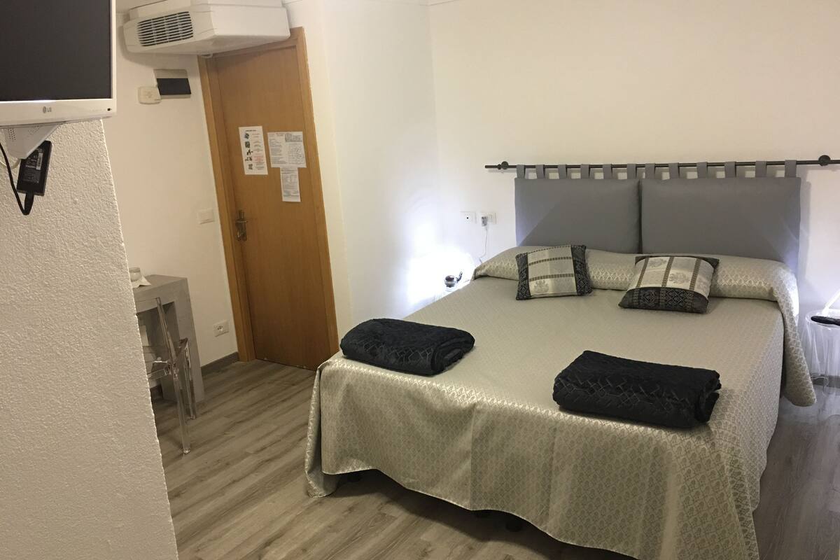 Una habitación de hotel con cama, una puerta de madera, aire acondicionado de pared y una mesita con lámpara.