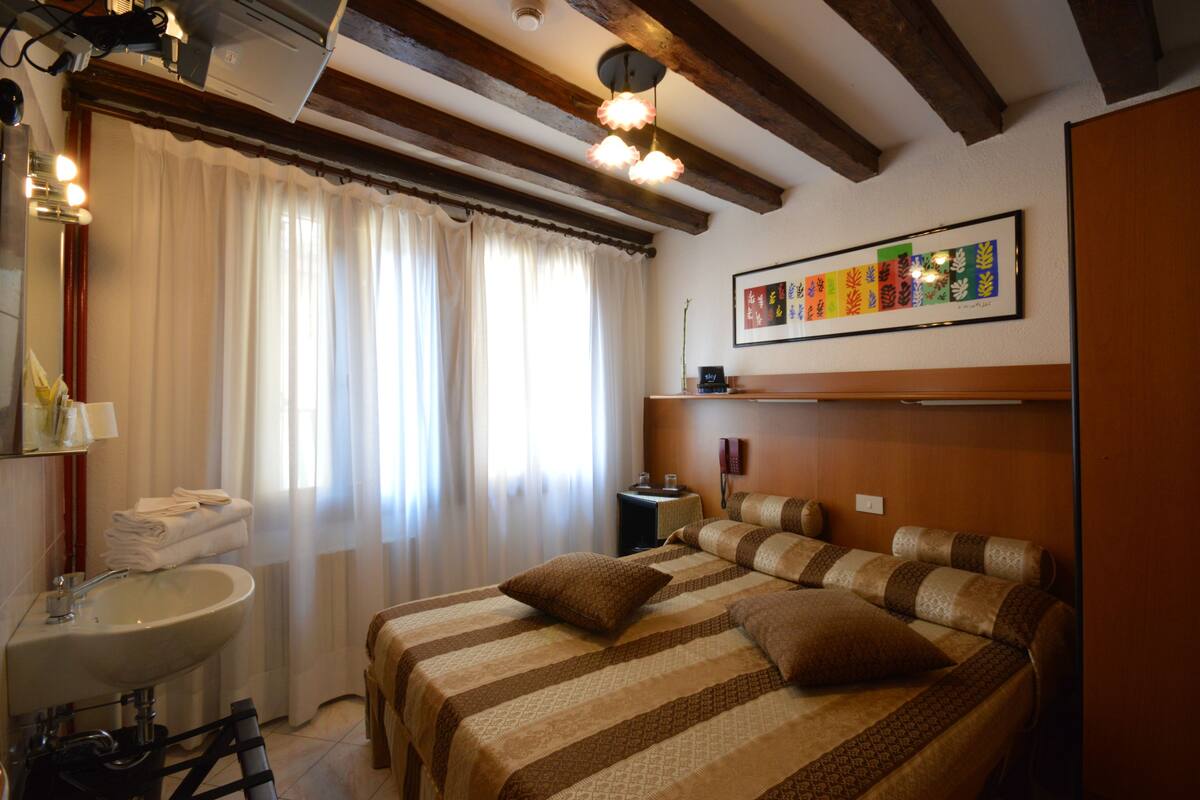Una habitación de hotel con una cama, una mesita de noche, una lámpara, una obra de arte enmarcada en la pared y un baño con lavabo e inodoro.