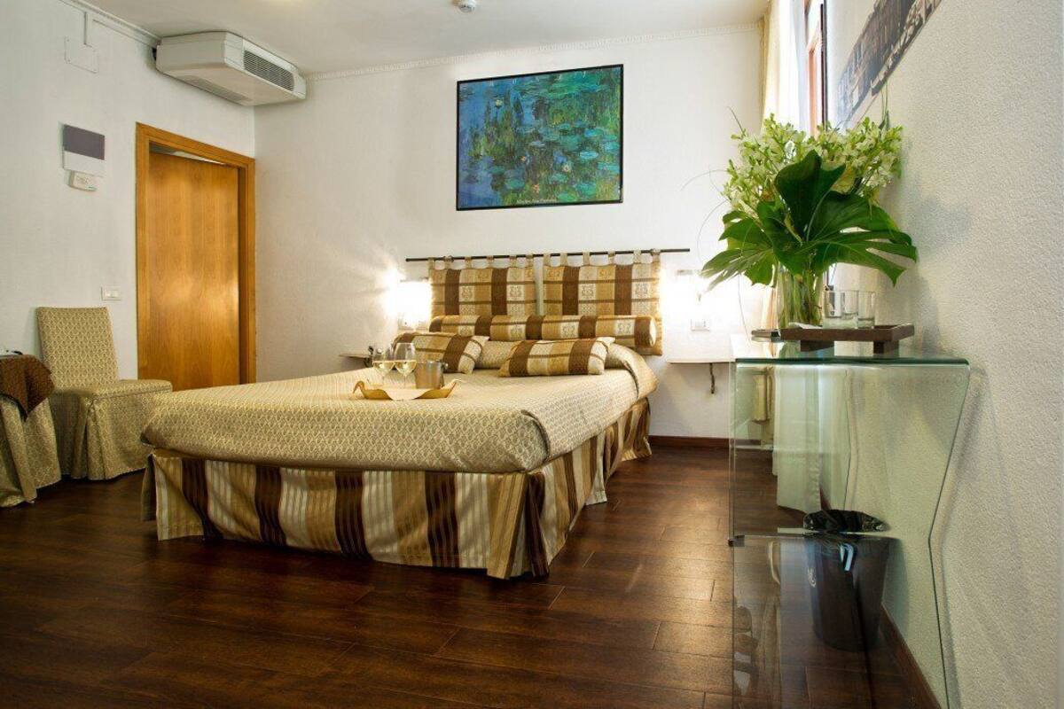 Habitación de hotel con una cama grande, una mesita de noche, una silla, una planta en maceta y un cuadro en la pared.