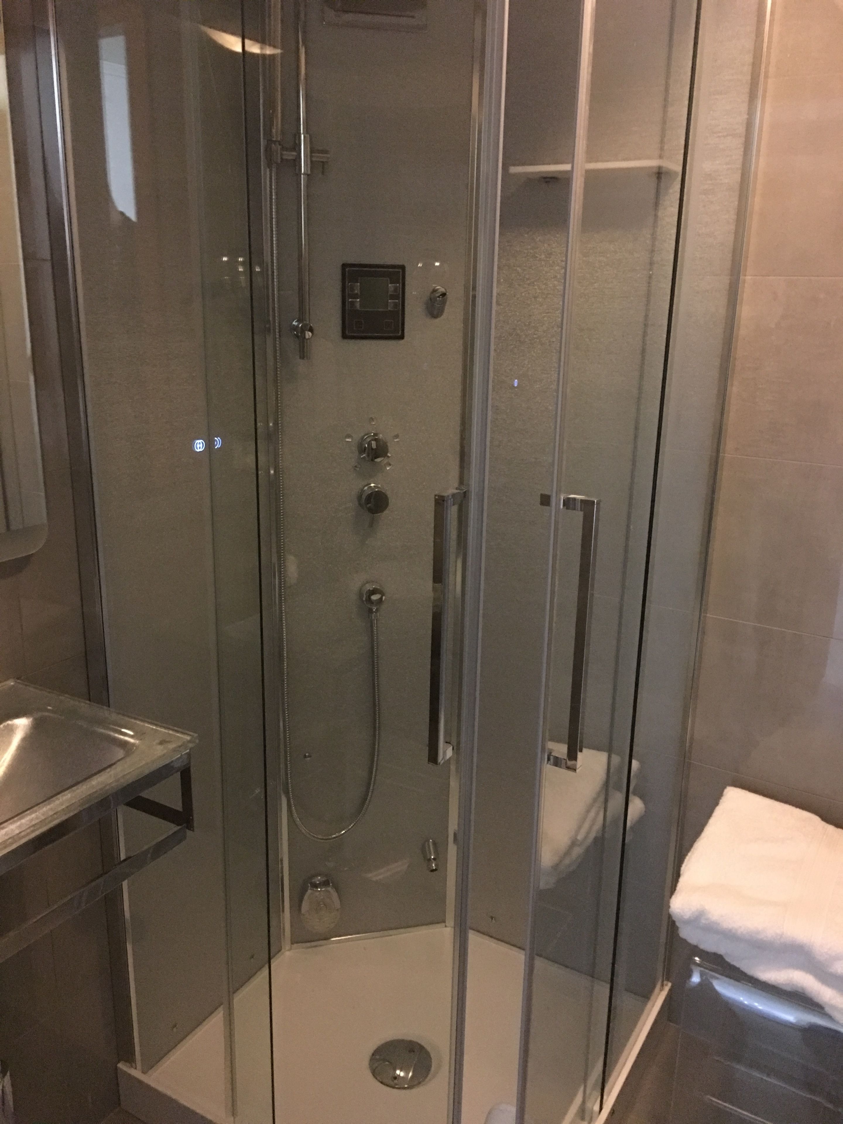A glass-enclosed shower stall with a handheld showerhead, a control panel, and a toilet visible through the glass.