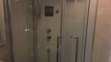 A glass-enclosed shower stall with a handheld showerhead, a control panel, and a toilet visible through the glass.