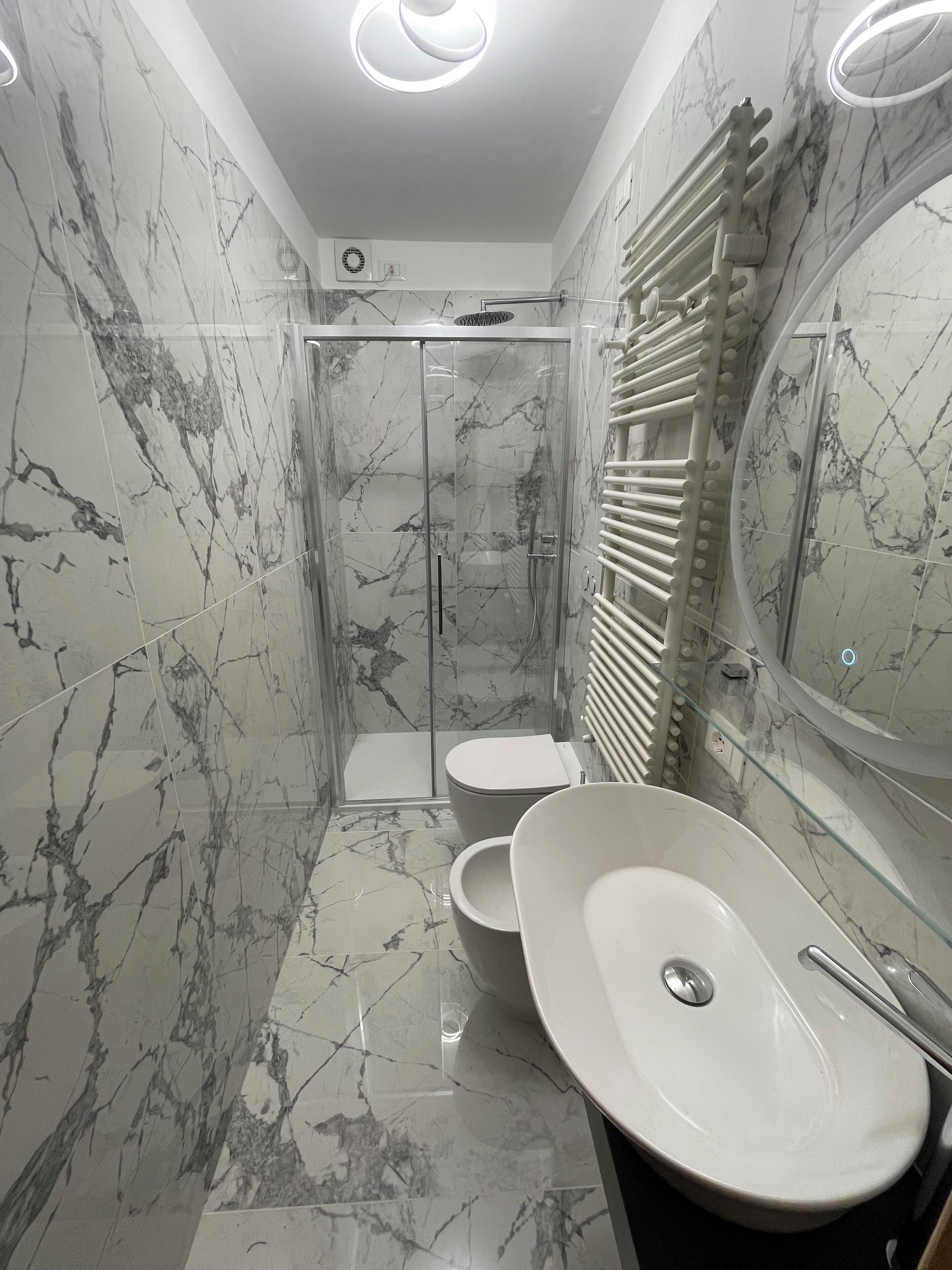 A modern bathroom with marble walls, a glass-enclosed shower, a toilet, and a double sink.