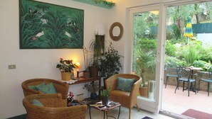 A cozy room with wicker furniture, a large green painting, and a view of a lush garden through a glass door. - Casa Popi (Malcesine)