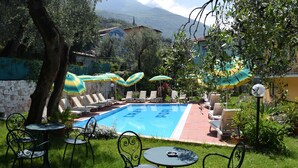 A pool area with lounge chairs and umbrellas, surrounded by greenery and a stone wall. - Casa Popi (Malcesine)