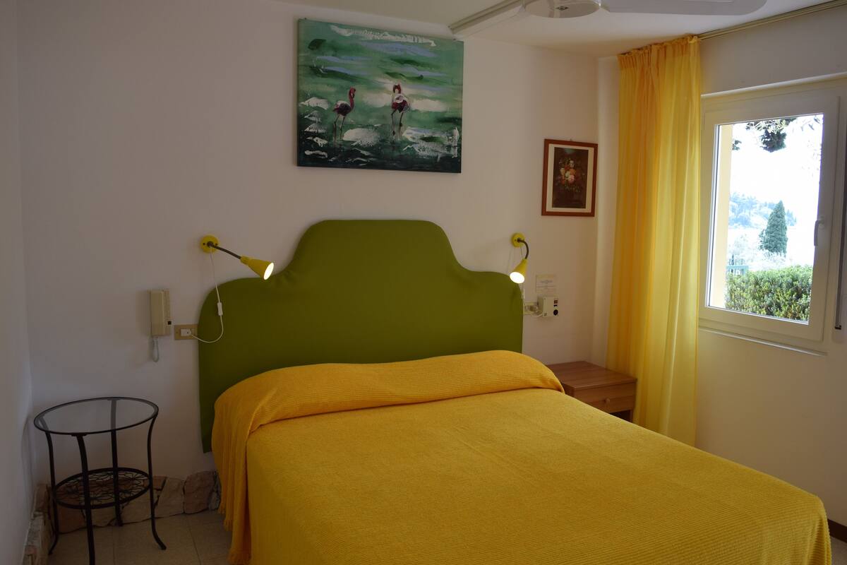 A room with a green headboard, a yellow bedspread, a painting of flamingos, a window with yellow curtains, and a small table with a lamp.