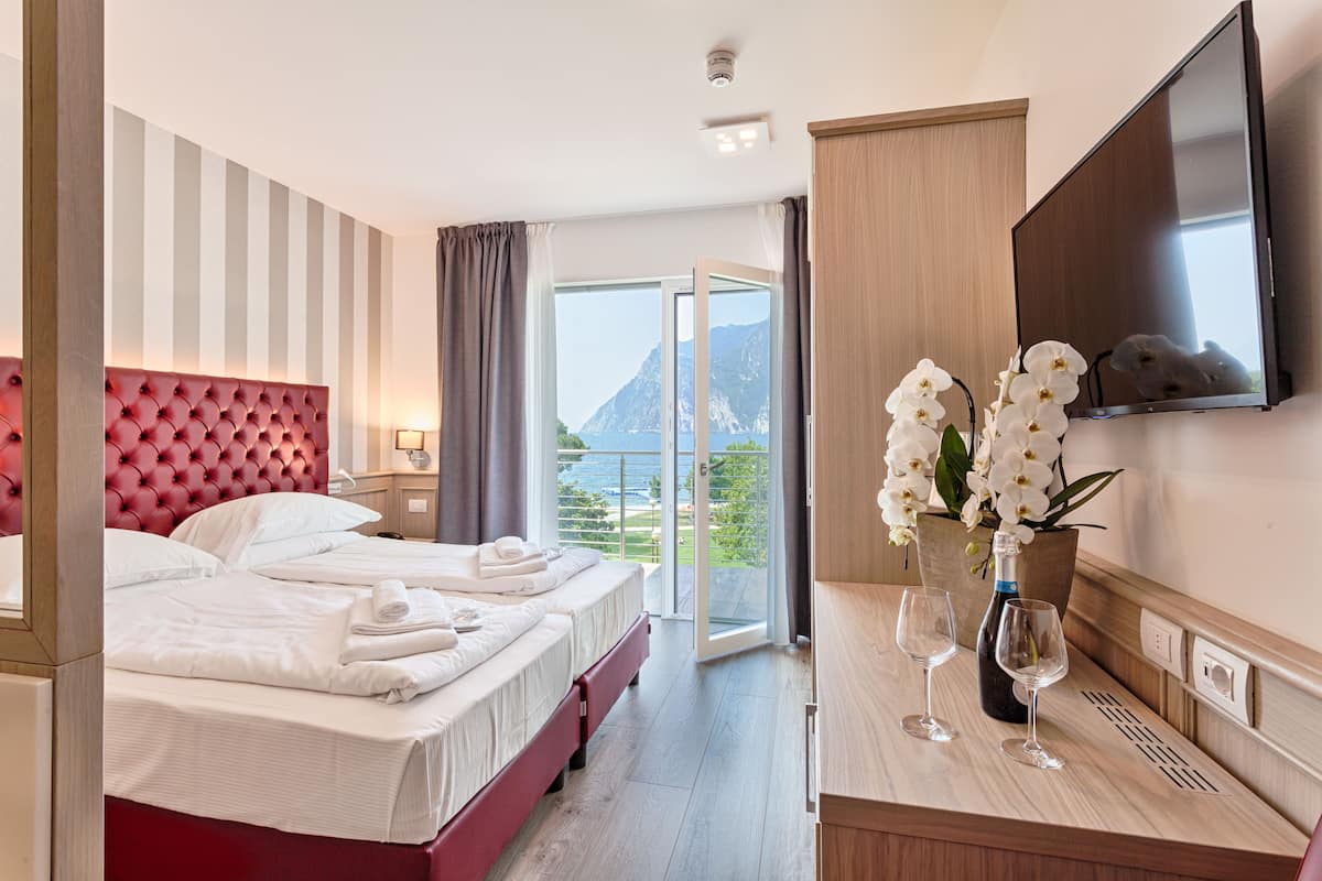 A hotel room with a red tufted headboard, two beds, a TV, a balcony with a mountain view, and a vase with white flowers.