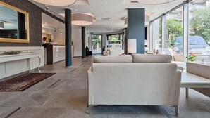 A modern hotel lobby with a reception desk, seating area, and large windows offering a view of the outdoors. - Bellariva (Riva del Garda)