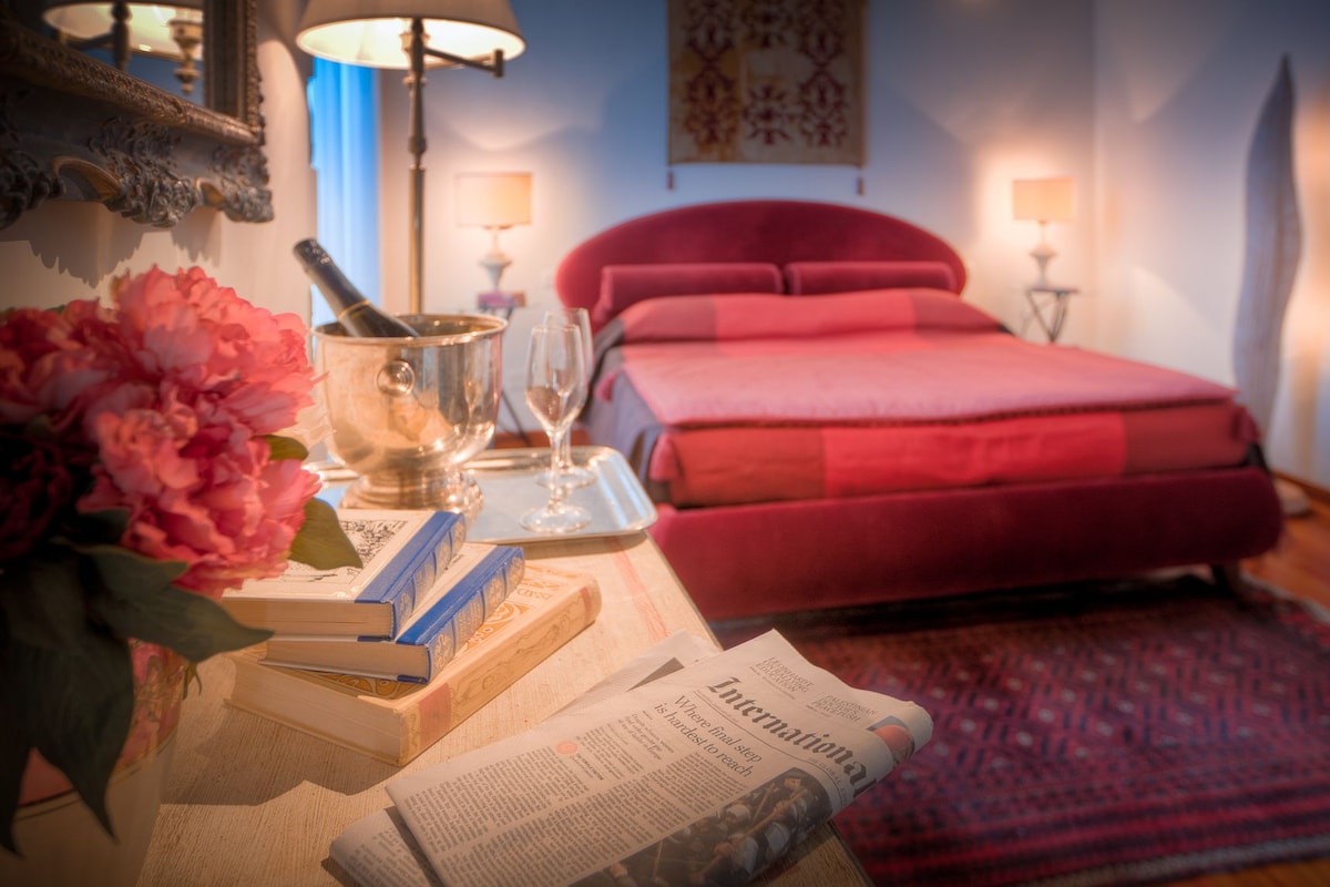 A bedroom with a red bed, a wooden bedside table with books, a lamp, and a floral arrangement.