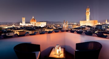 A rooftop terrace with wicker chairs and a table, overlooking a cityscape with historic buildings and a prominent tower.