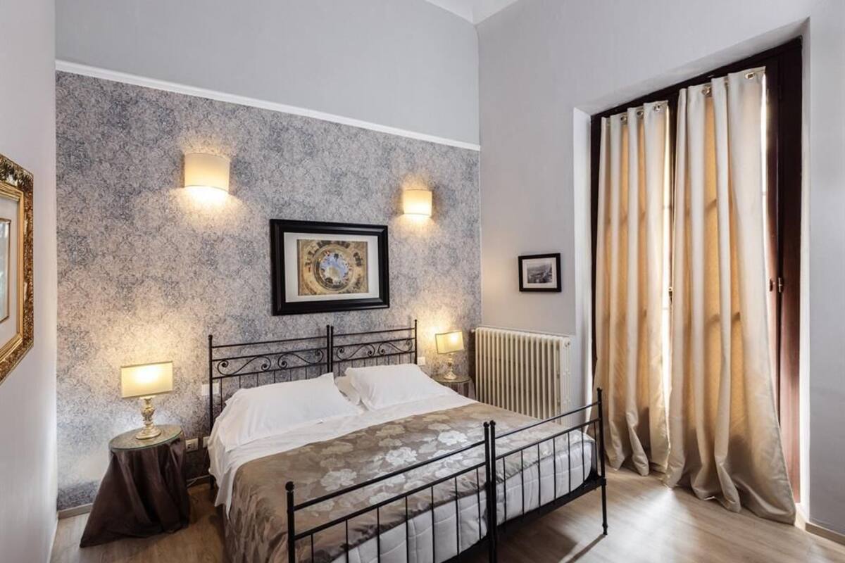A bedroom with a bed, two wall-mounted lamps, a radiator, and a window with curtains.