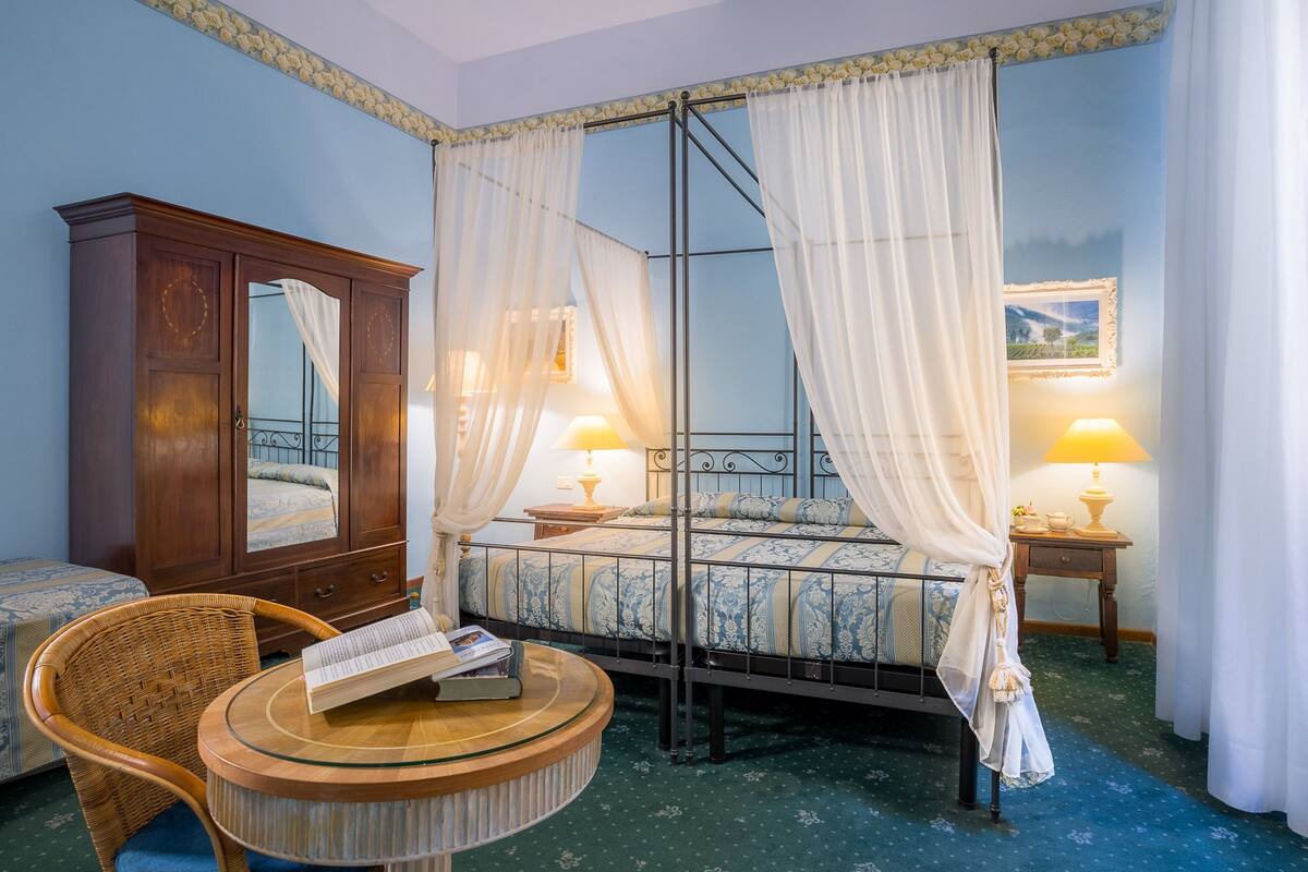 A bedroom with a four-poster bed, a wooden wardrobe, a wicker chair, and a round table with books.