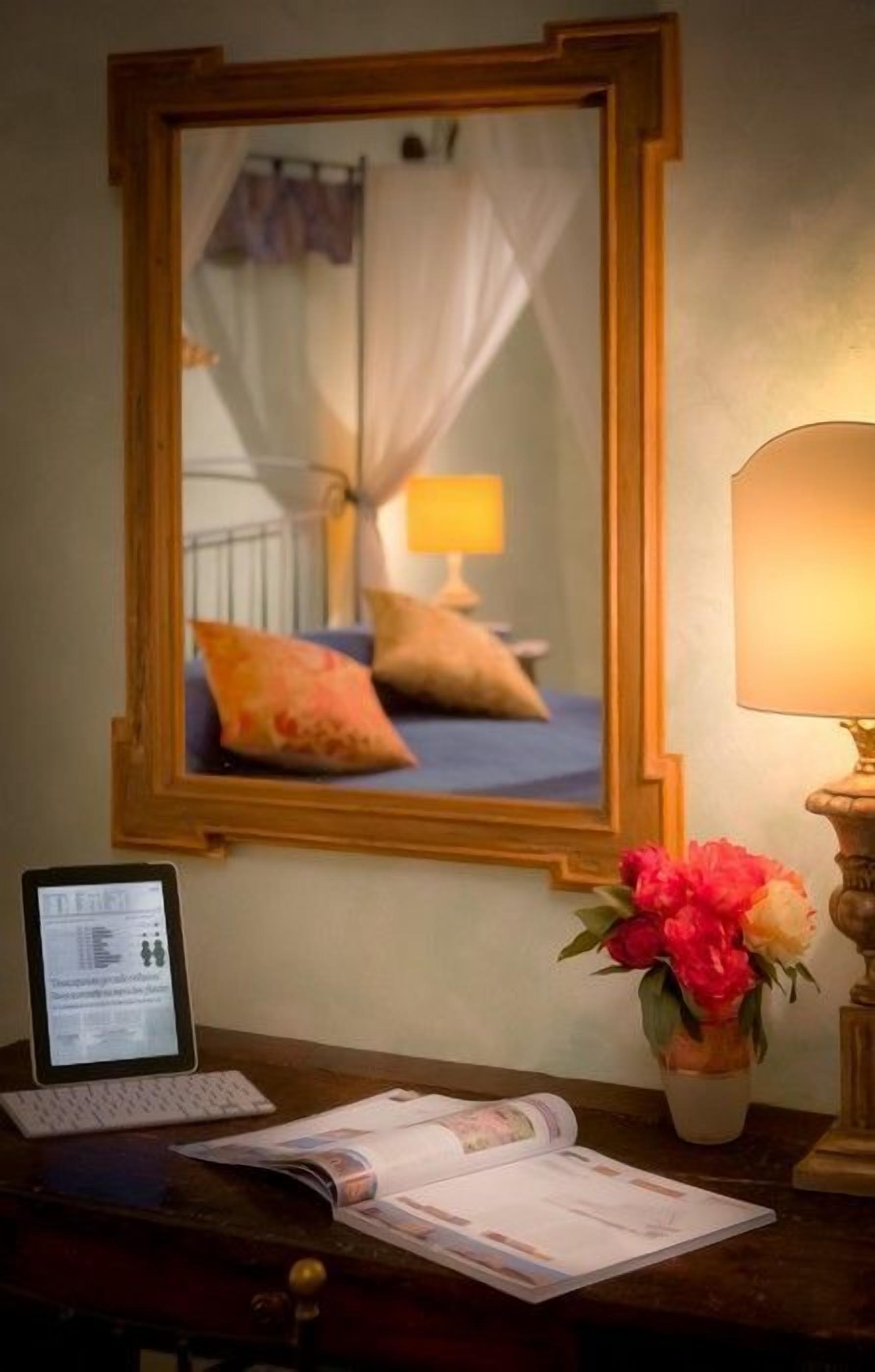 A hotel room with a bed, a bedside lamp, a laptop, a vase with flowers, and a mirror reflecting the room.
